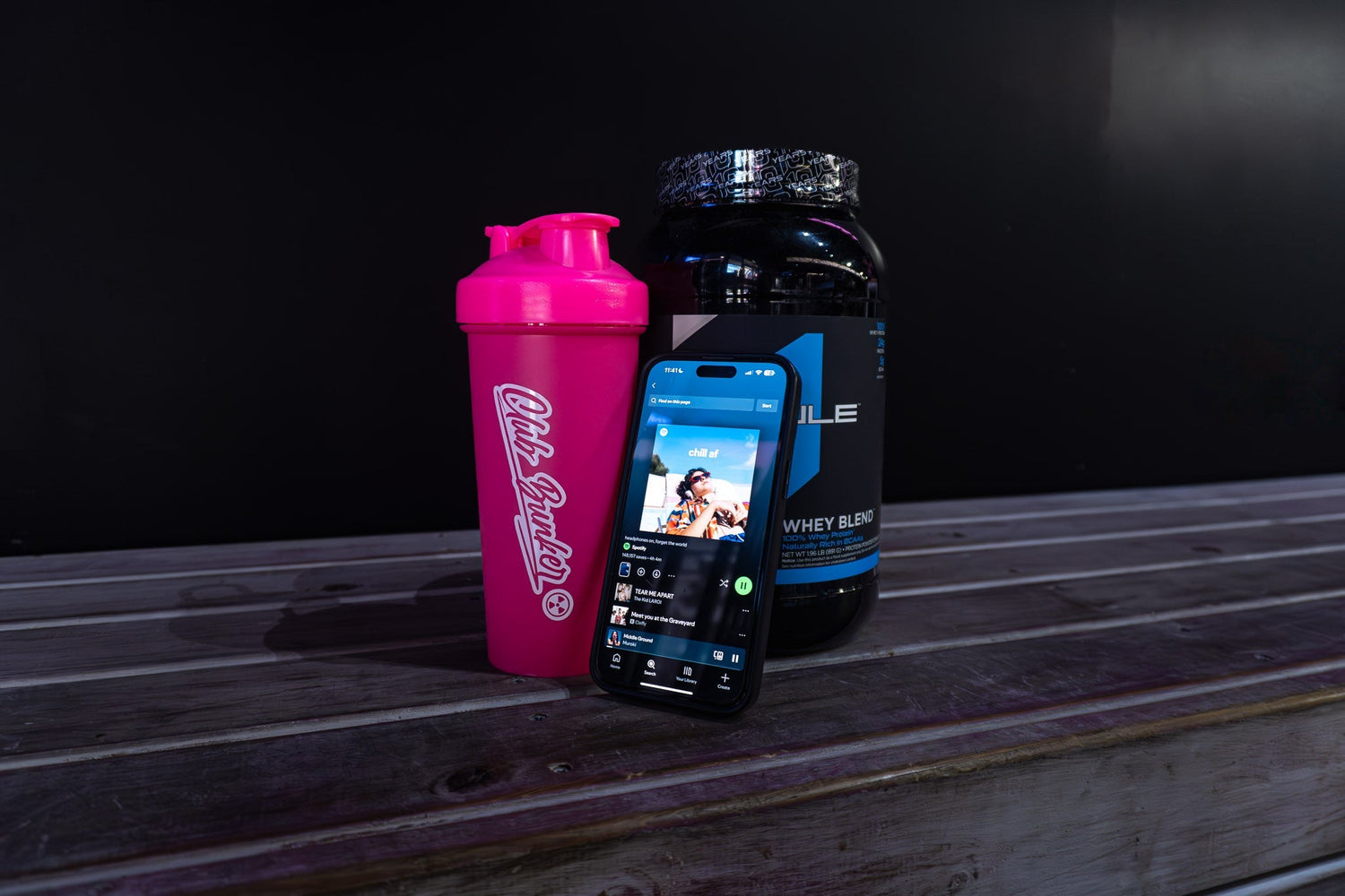Club Bunker pink shaker and Rule1 protein beside a phone playing Spotify on a timber bench with dark background.