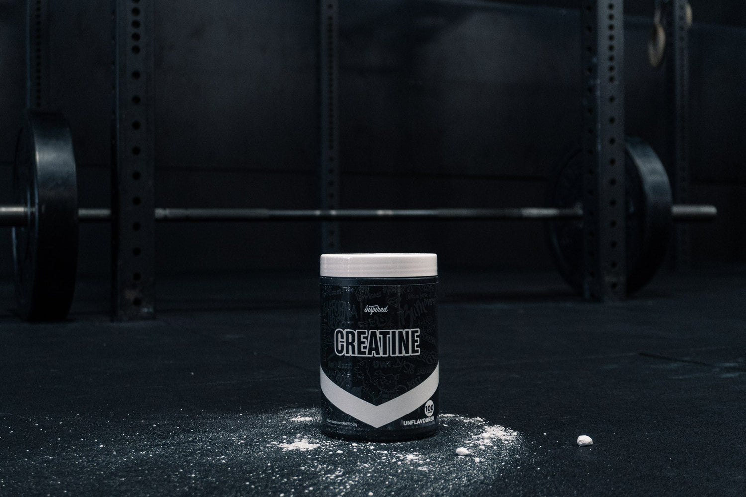 Inspired Creatine tub surrounded by chalk in front of loaded barbell and squat rack in a dark gym setting.